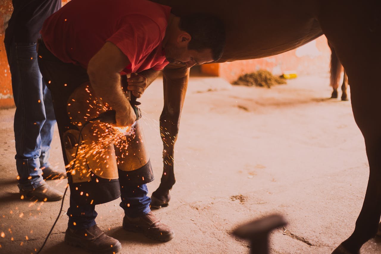 A farrier shoeing a horse in an indoor stable with flying sparks from the equipment.